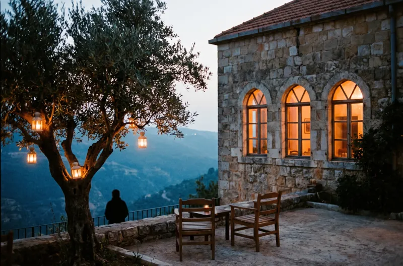 An old stone house with three arched windows glowing at blue hour, an olive tree with lanterns on the terrace.