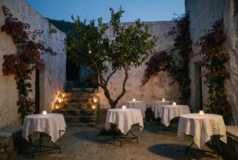A whitewashed courtyard at dusk with a lemon tree, small round tables in linen and candles on the stairs.